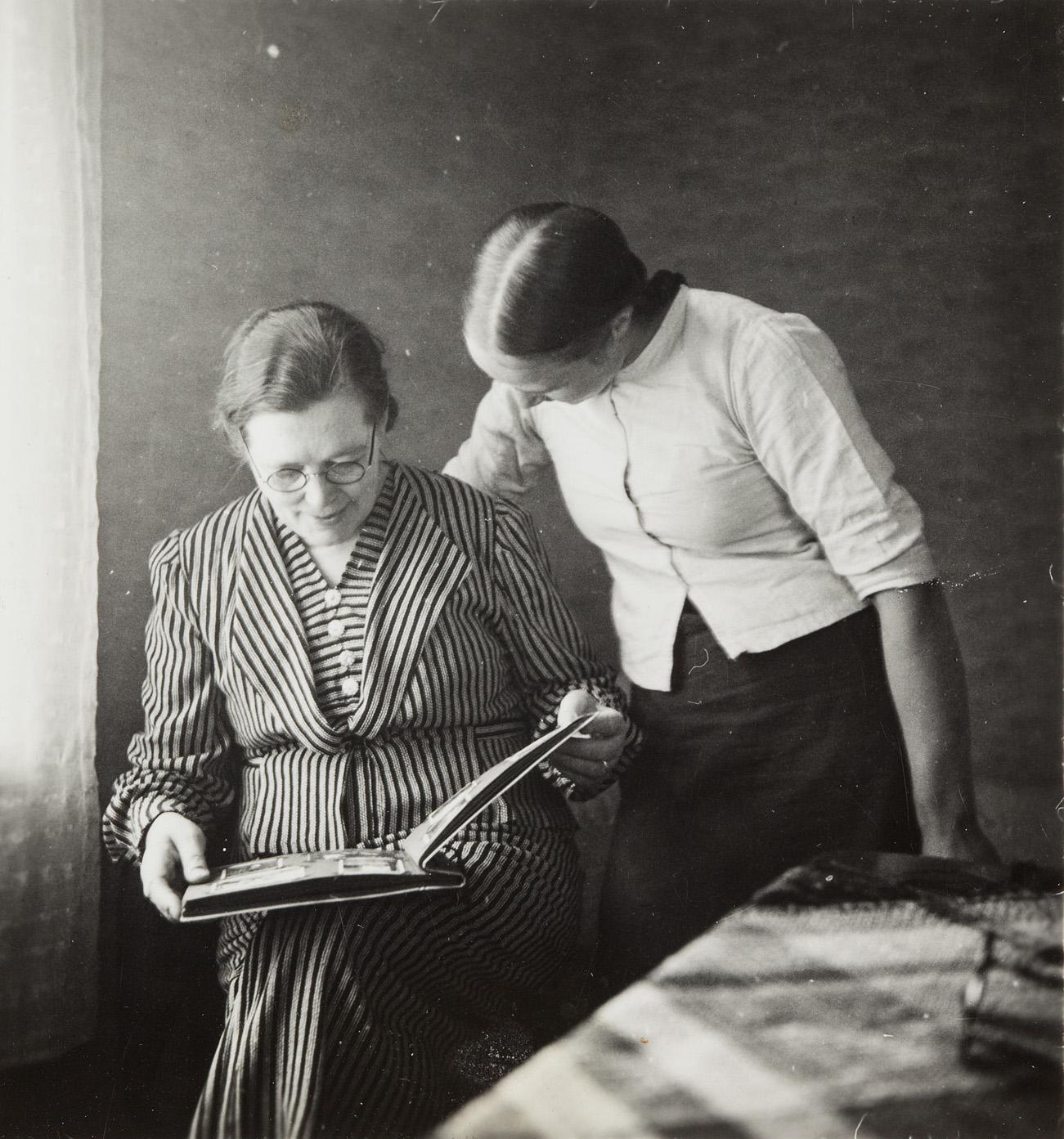 Kuva: Joseph Hart, 1930-1950. Kaksi naista tarkastelemassa valokuva-albumia, vas. Olga Hart. Vasemmalla on ikkuna, josta lankeaa päivänvaloa. D1973:4/1908 / Suomen valokuvataiteen museon kokoelma